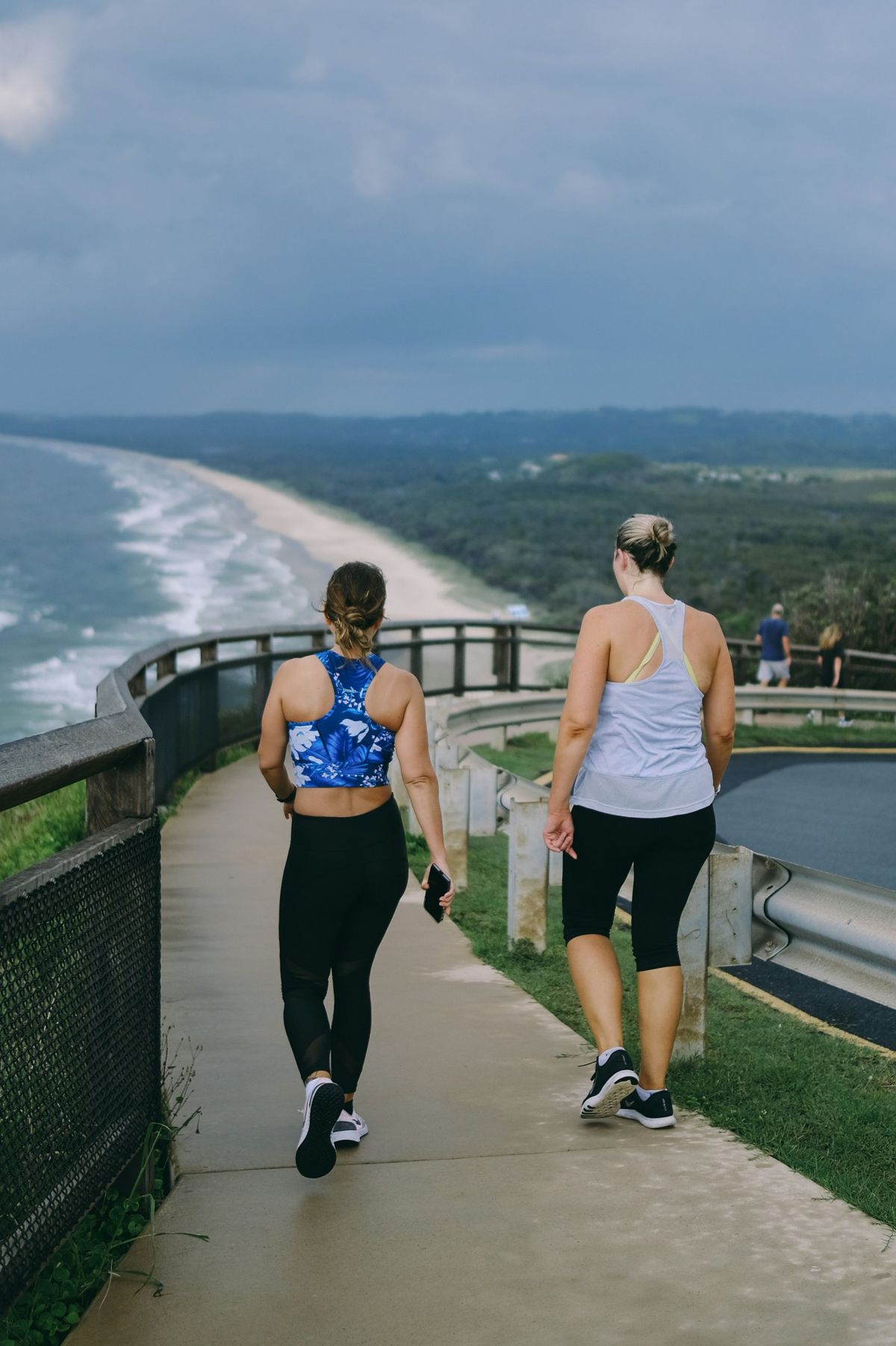 Two women walking together on a coastal lookout path above a long beach